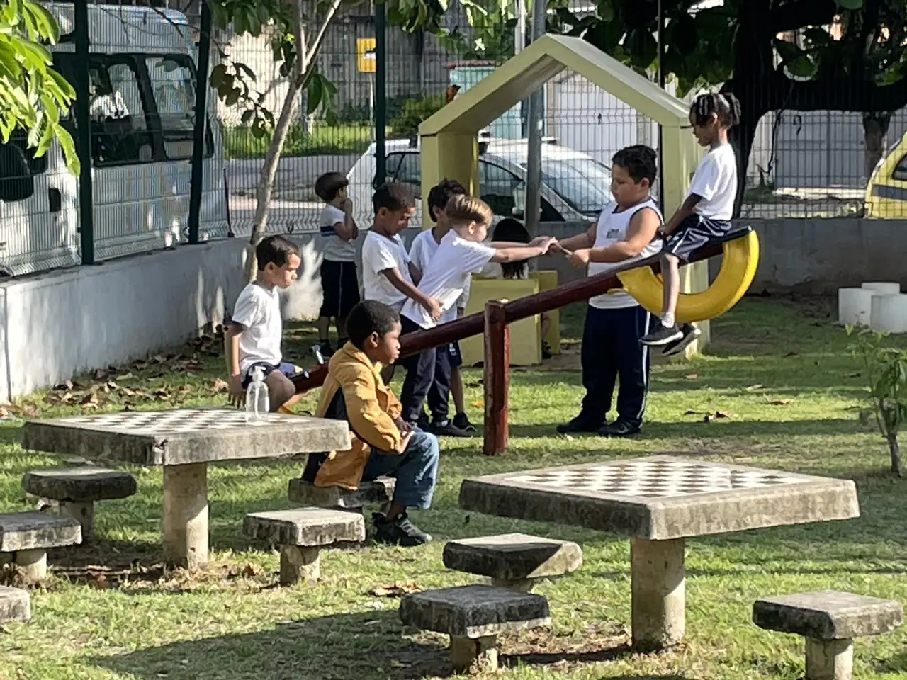Grupo de crianças brincando juntas em uma gangorra, ao lado de mesas de pedra em uma praça