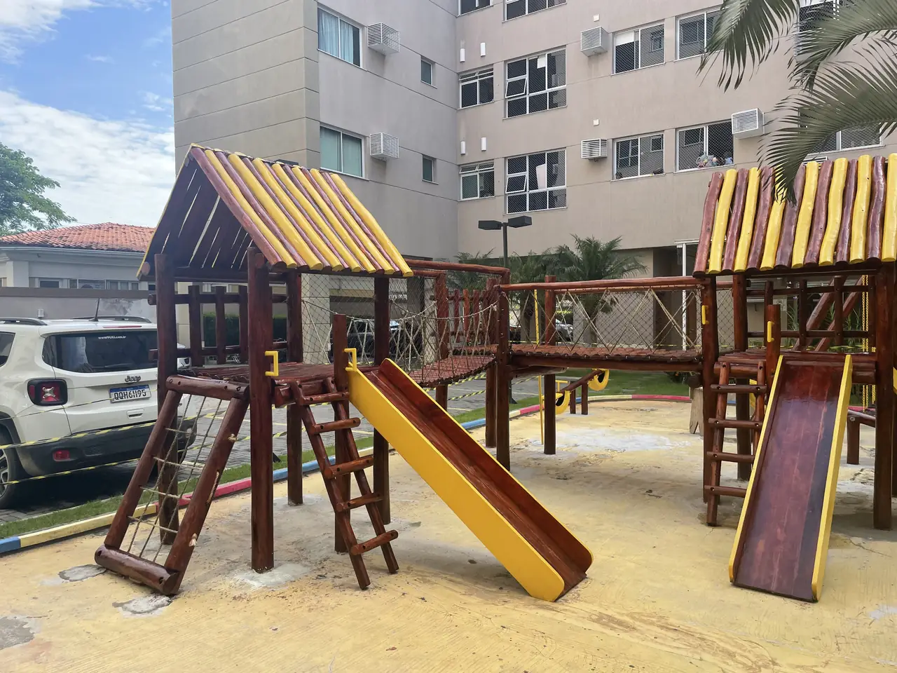 Playground de madeira com torres e escorregadores sobre piso de areia em condomínio